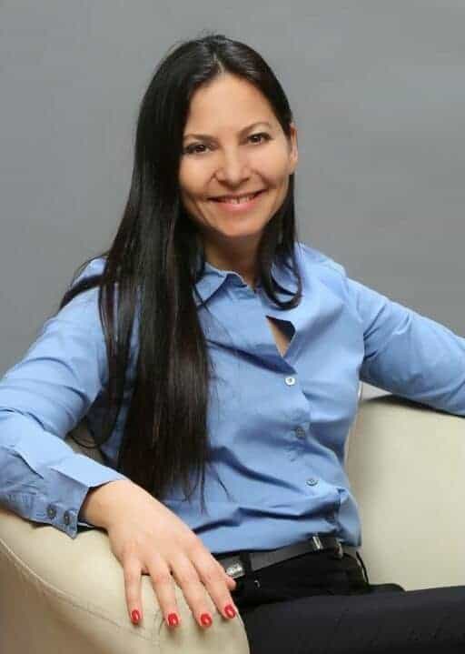 אישה עם שיער ארוך וכהה, לבושה בחולצה מכופתרת בצבע תכלת, יושבת ומחייכת למצלמה על רקע אפור אחיד. | גליה גביש מטפלת LI-CBT ילדים, נוער ומבוגרים ומדריכת הורים